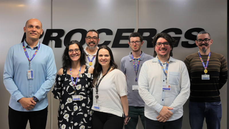 Foto mostra sete integrantes da da equipe posados em frente ao logo da Procergs